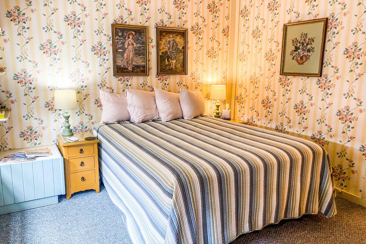 Une chambre à coucher avec une couvre-lit à rayures, un papier peint à motifs floraux, deux tableaux encadrés, une lampe de chevet et une table de chevet avec une lampe et une boîte à mouchoirs.