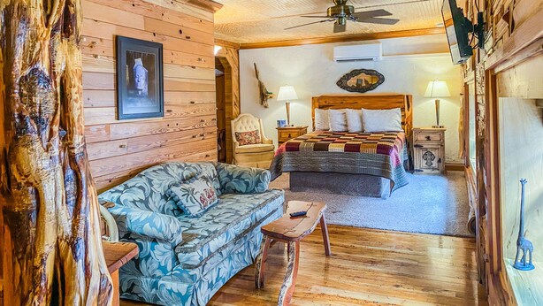 A log cabin-style bedroom with a wooden ceiling, a bed with a quilt, a sofa, and a small table. - Inn at the Art Center (San Angelo)