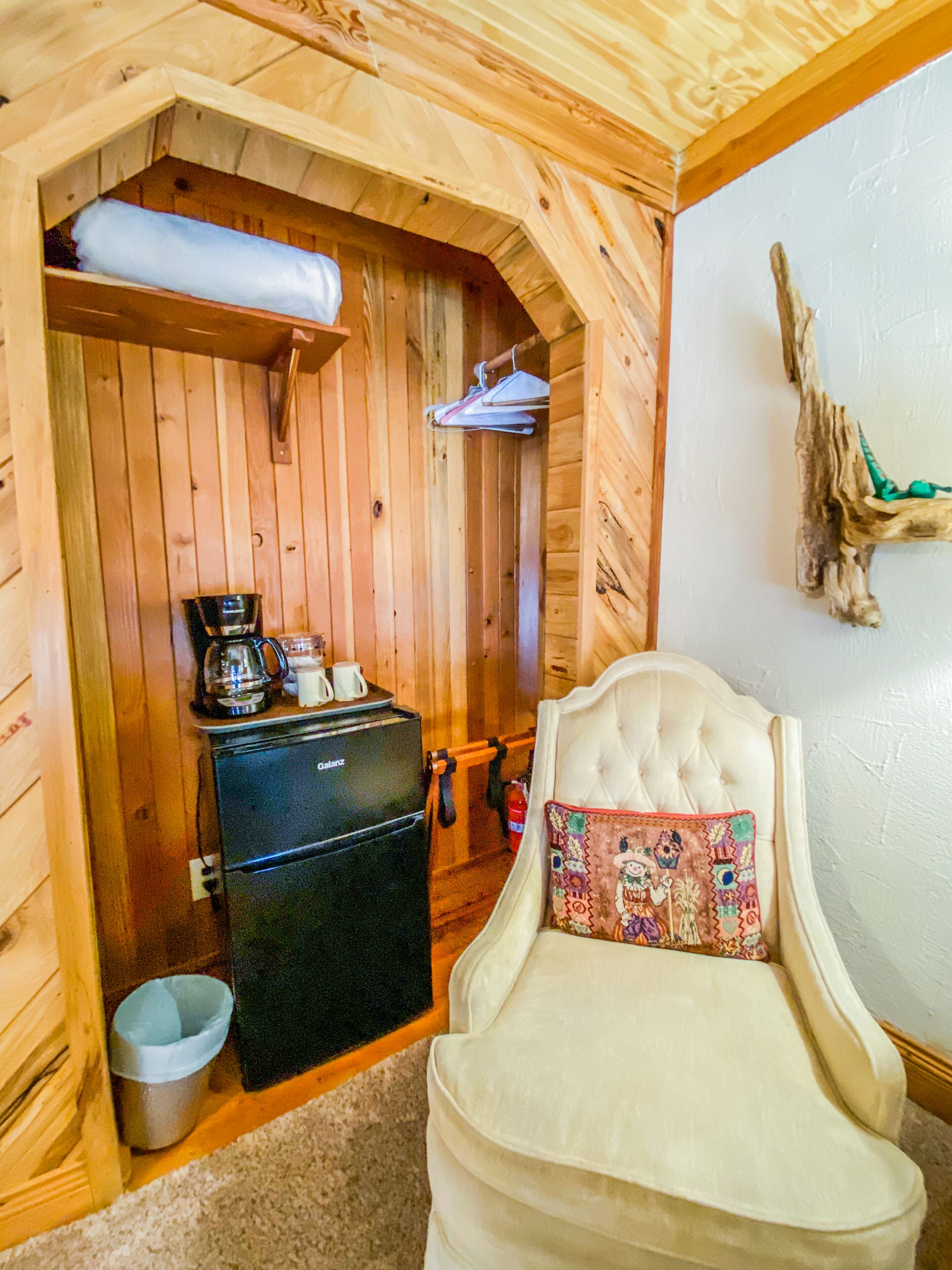 Ein gemütliches Zimmer mit Holzwänden, einem weißen Polsterstuhl mit dekorativer Kissen und einem kleinen schwarzen Kühlschrank, einer Kaffeemaschine sowie einem Kleiderhaken.