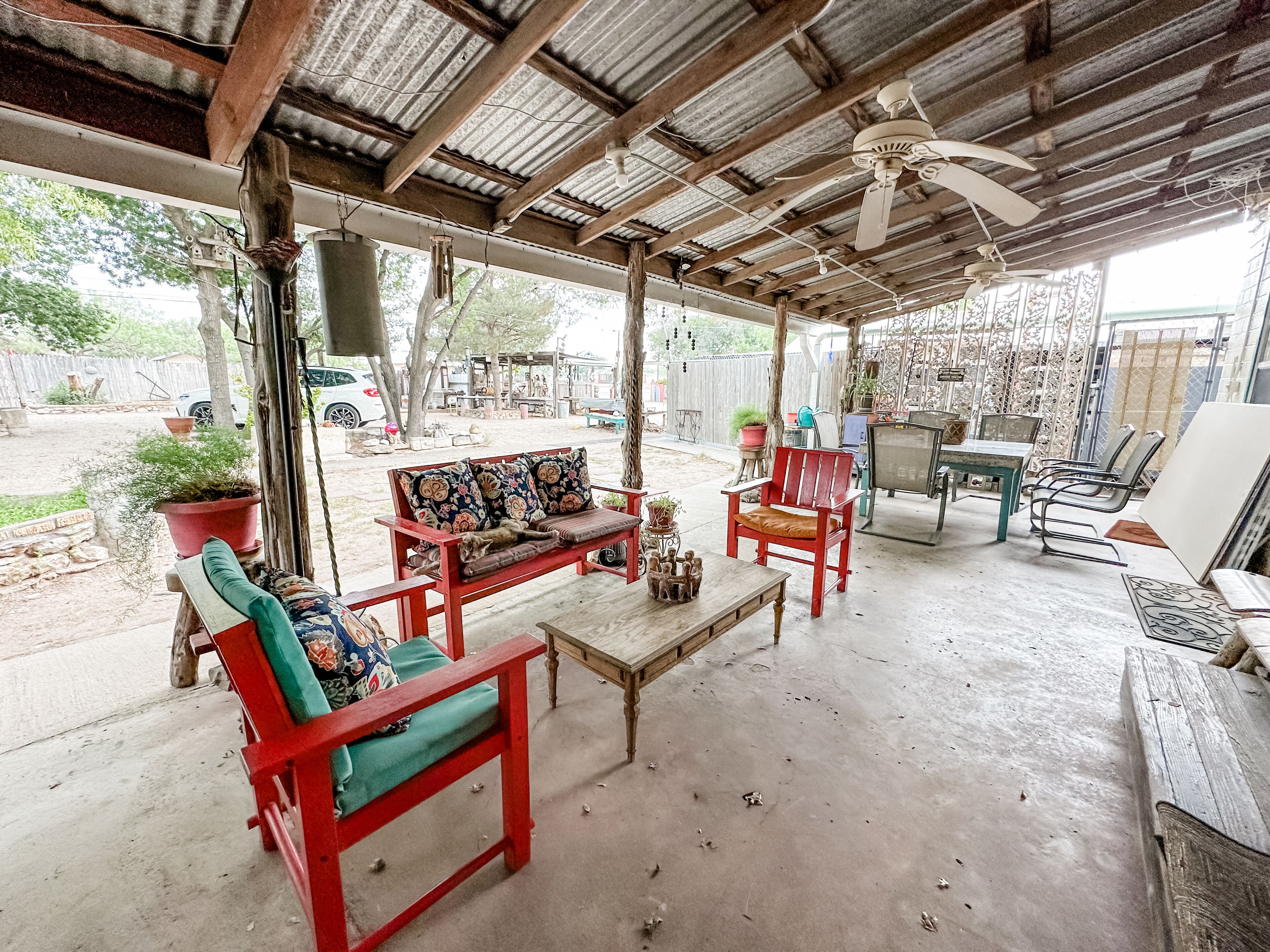A covered outdoor seating area with a wooden bench, a red chair, and a sofa with floral cushions.