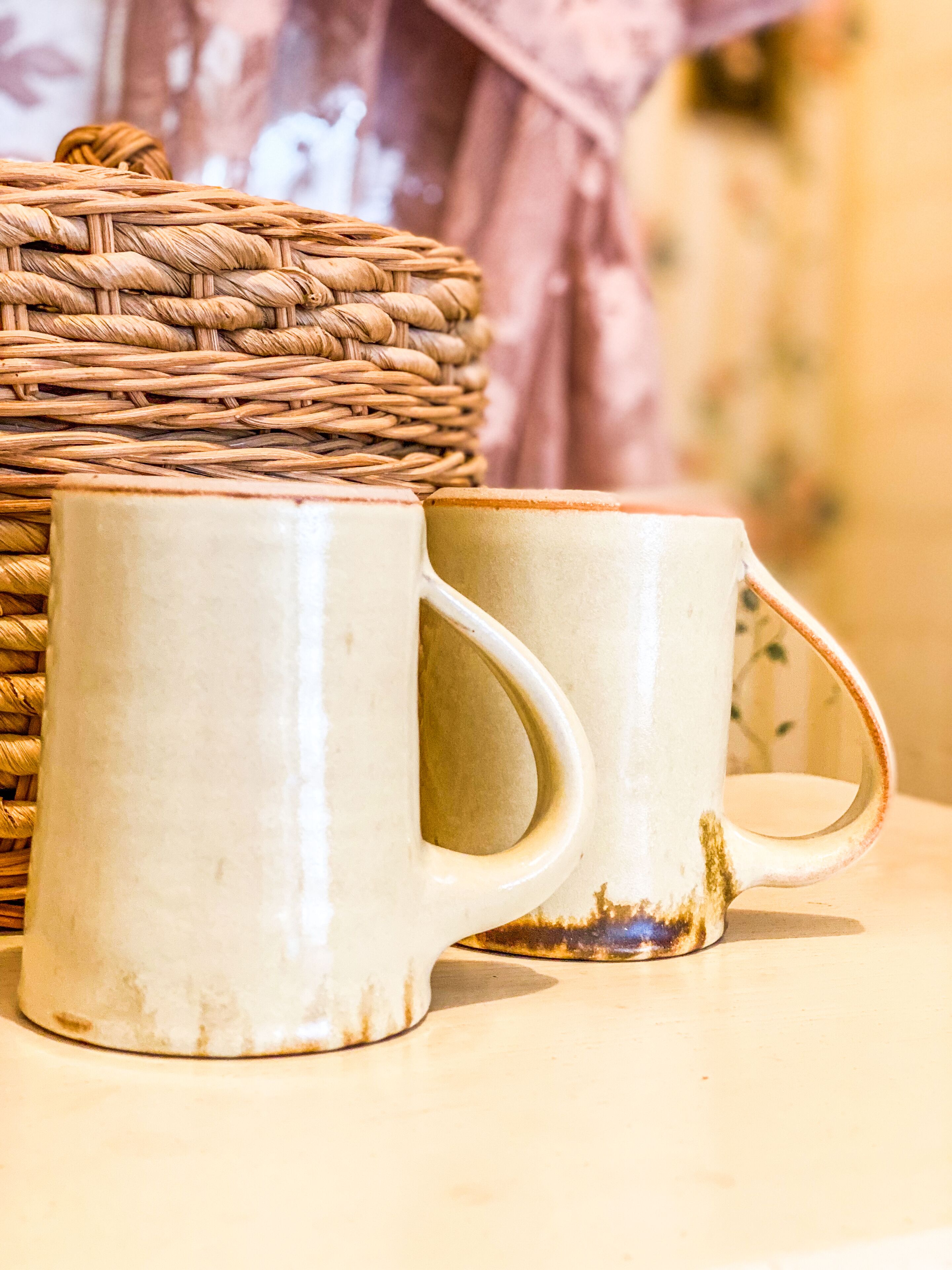 Deux tasses en céramique blanche, dotées d’anses, posées sur une surface, avec un panier en osier en arrière-plan.