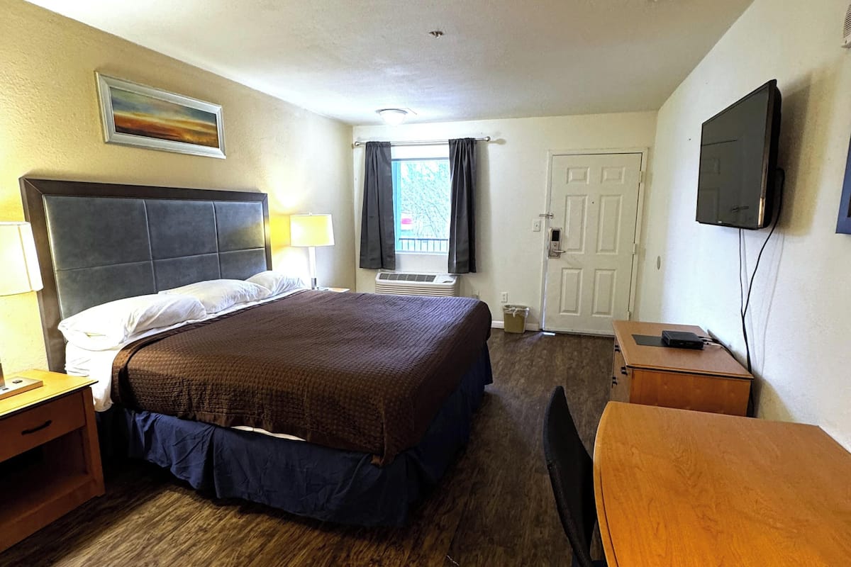 A hotel room with a bed, a desk, a TV, a window with curtains, a door, and a painting on the wall.