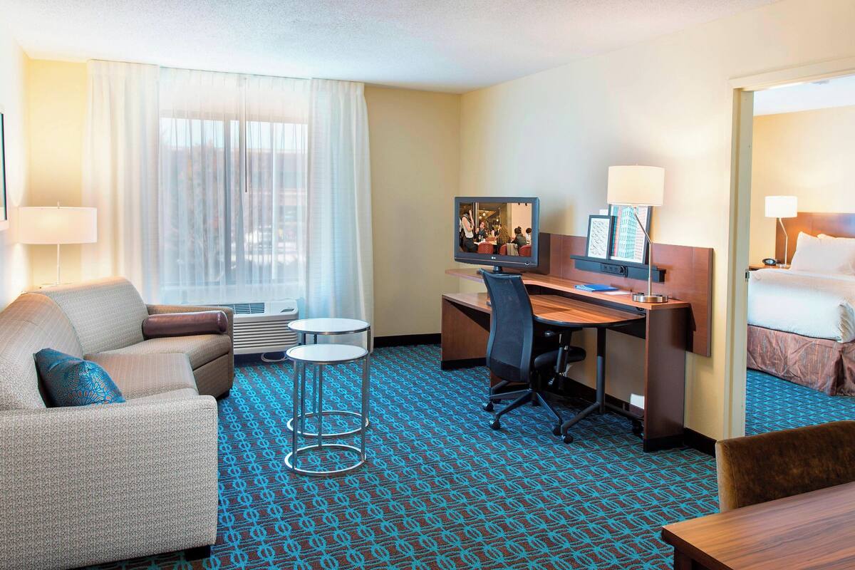 Habitación de hotel con sofá, escritorio, silla, cama, televisor y ventana con cortinas.