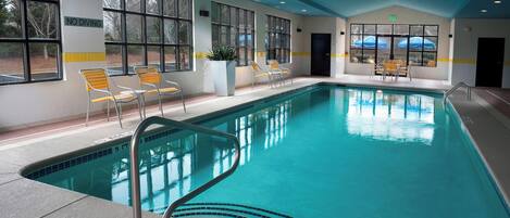 Una piscina interior con agua azul cristalina, sillas amarillas y blancas, y ventanas amplias.