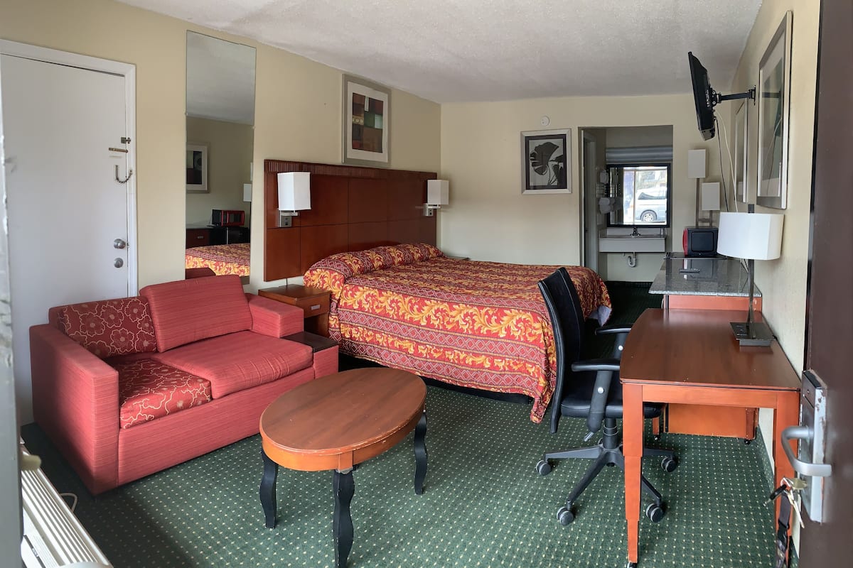 A hotel room with a bed, a red sofa, a wooden desk, a chair, a small table, and a mirror.