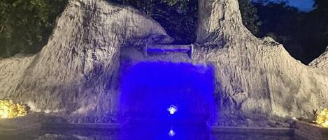 A nighttime outdoor water feature with a blue-lit pool, surrounded by rock formations and trees.