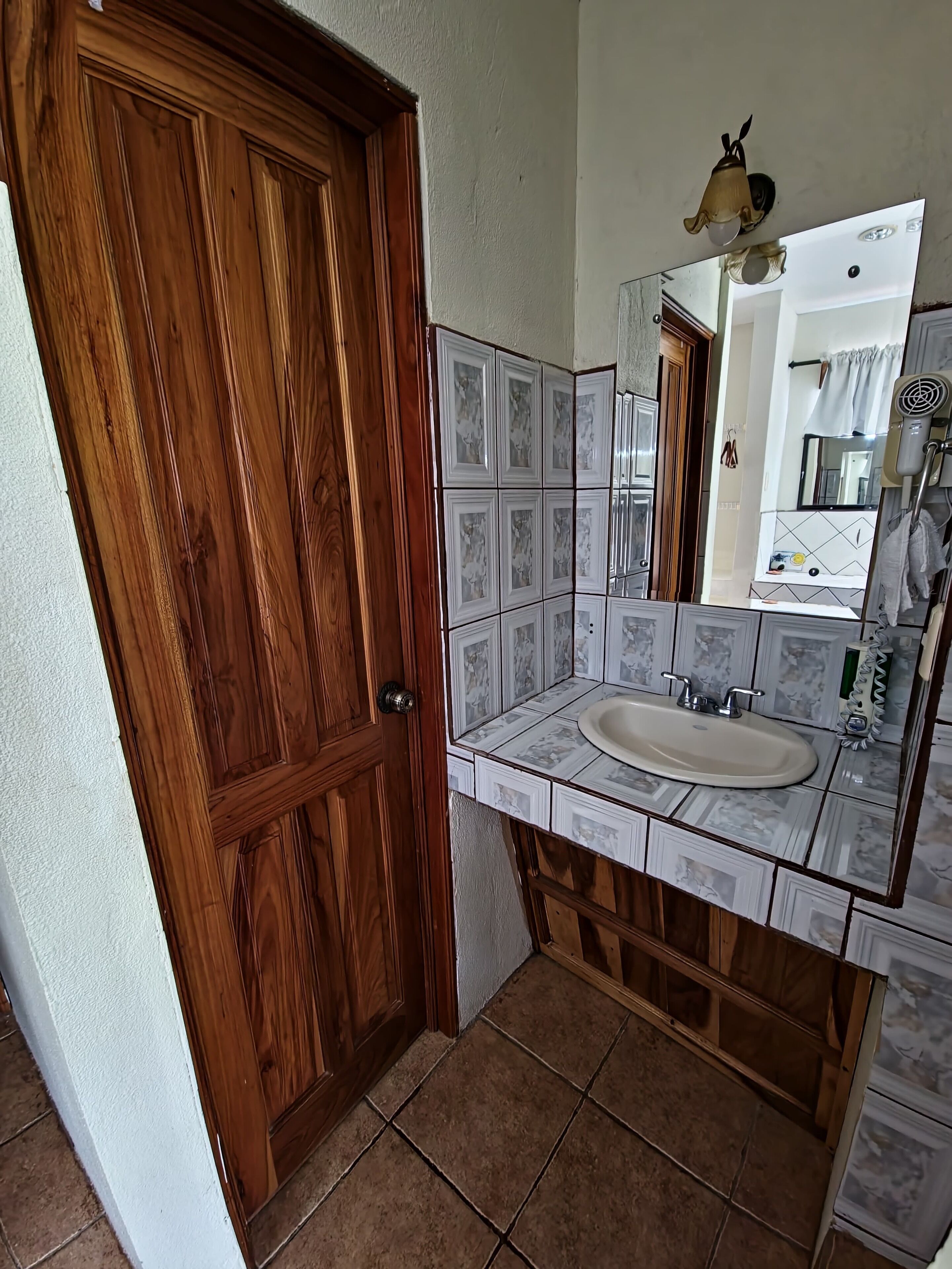 Casa de banho com porta de madeira, pia com espelho e azulejos estampados.