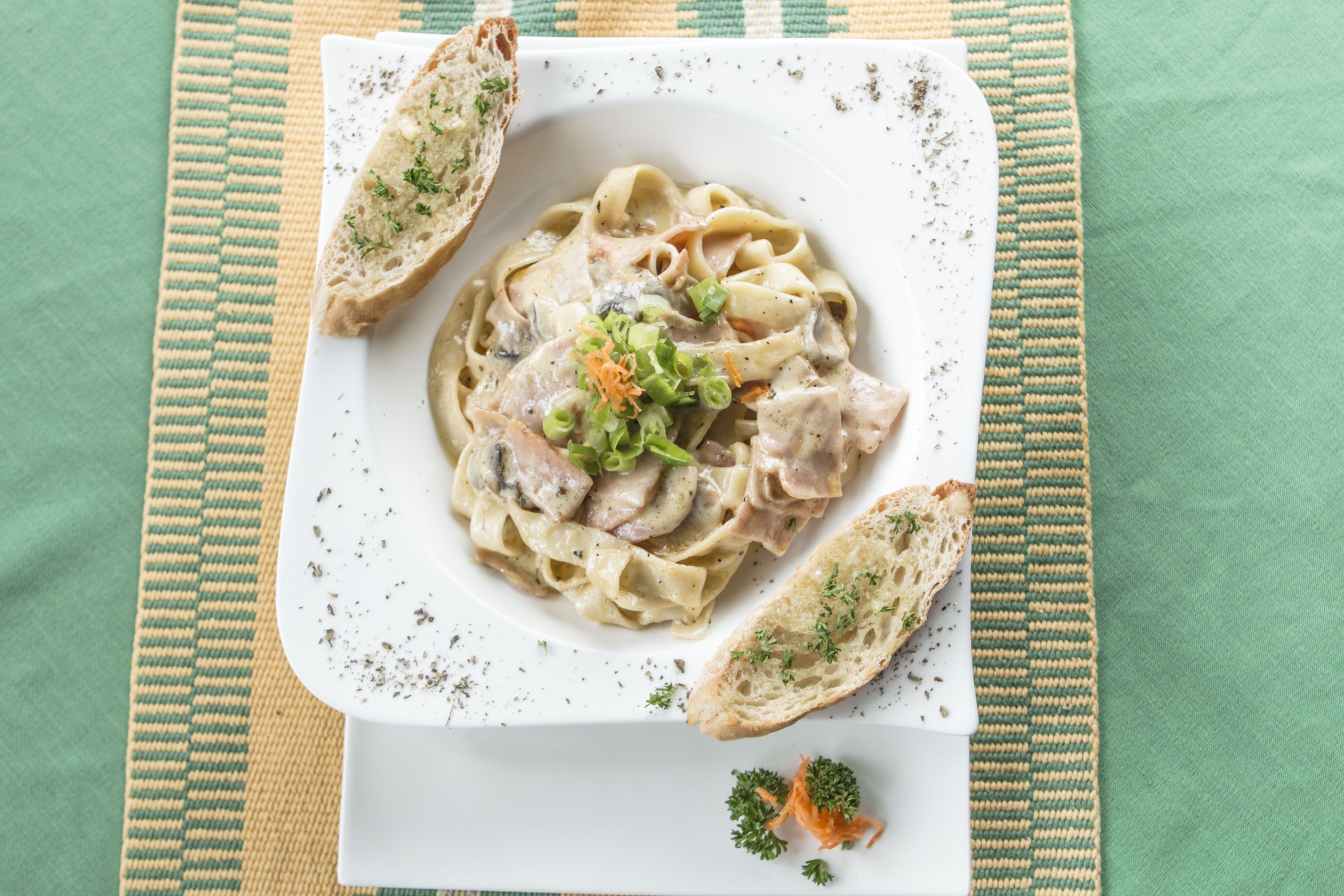 Une assiette de pâtes nappées d’une sauce crémeuse, garnie d’herbes fraîches, et accompagnée de tranches de pain sur un set de table en fibres tressées.