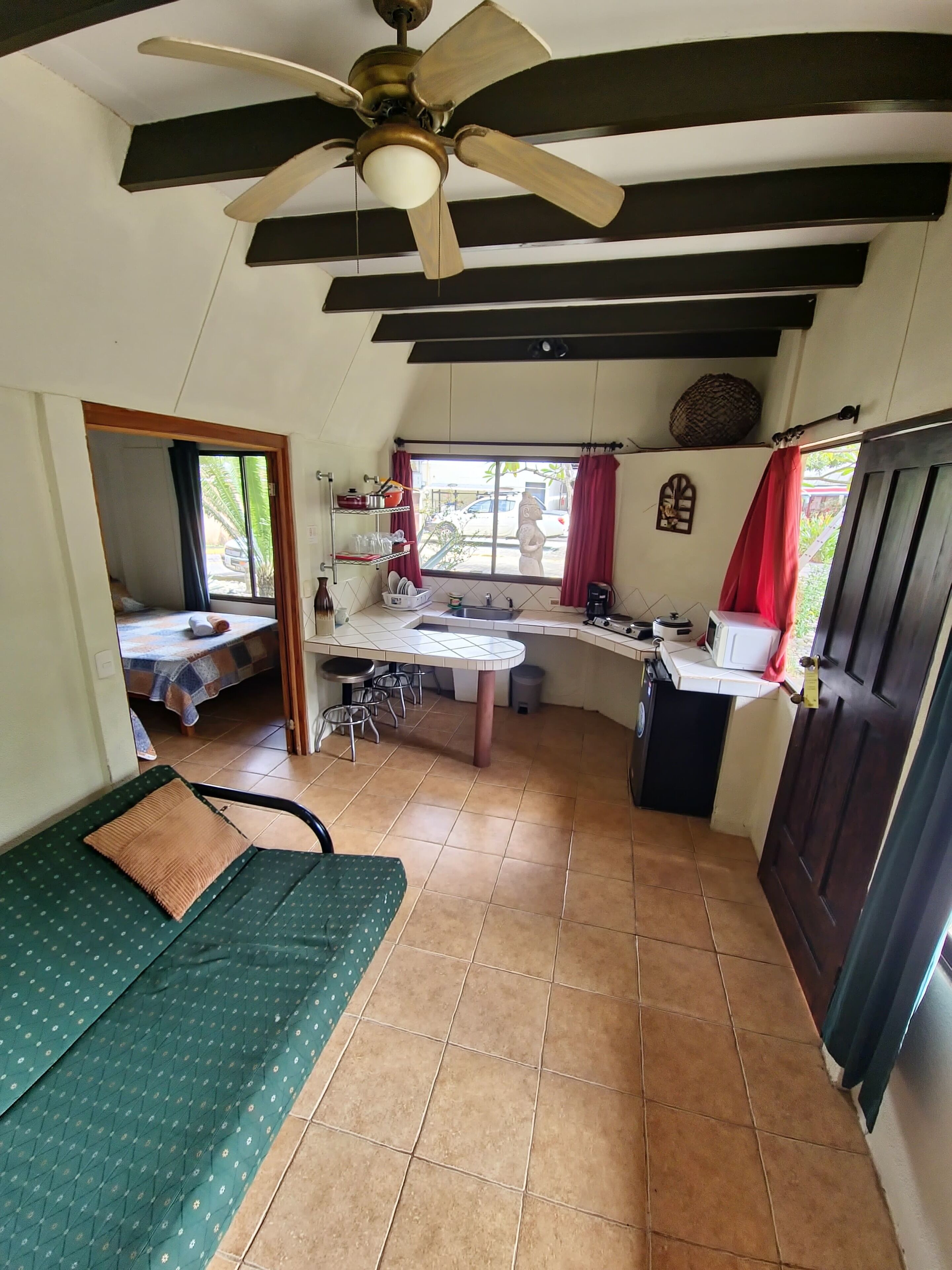 A compact kitchen with a dining area, a ceiling fan, and a window with red curtains.