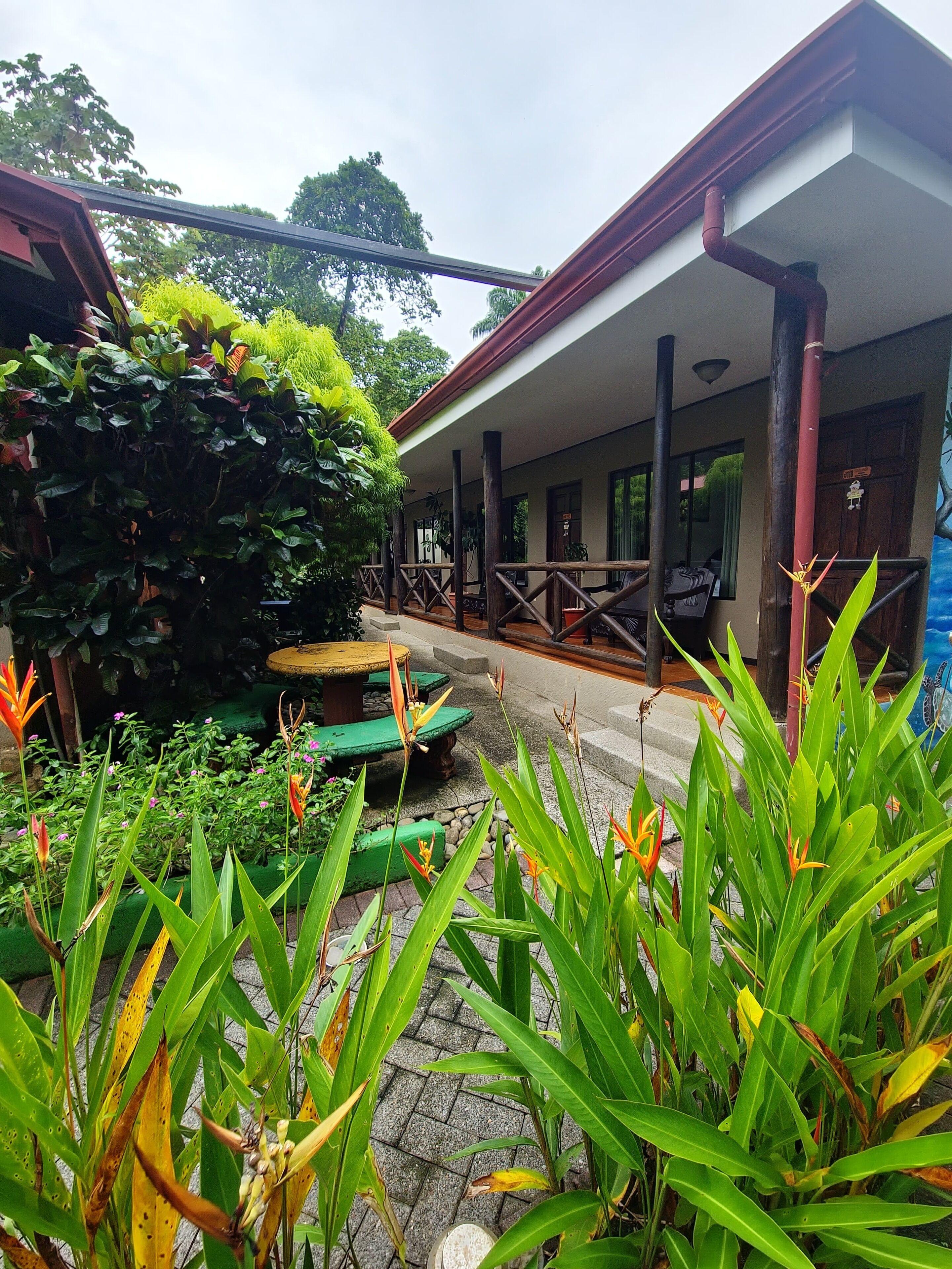 A covered outdoor area with a wooden bench, potted plants, and a pathway leading to a building with a red roof.