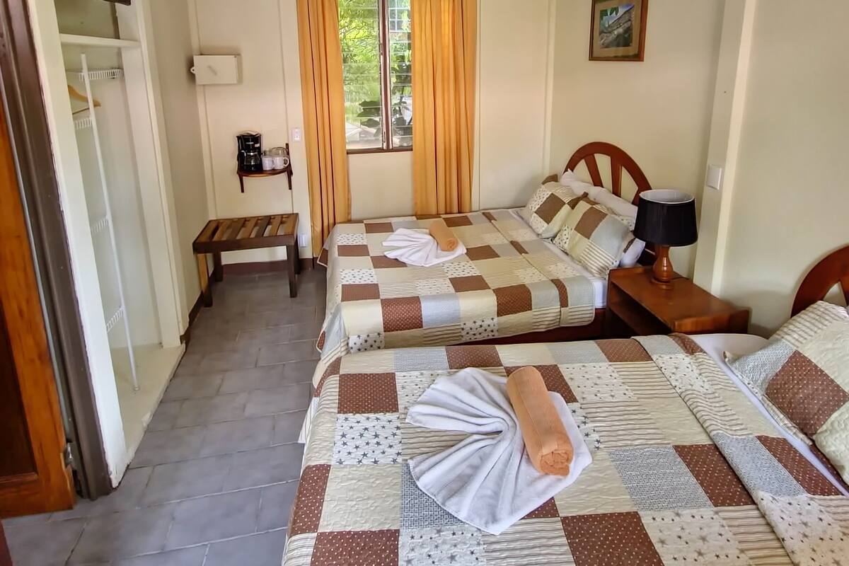 A hotel room with two beds, a ceiling fan, a window with curtains, a bedside table with a lamp, and a small bench.