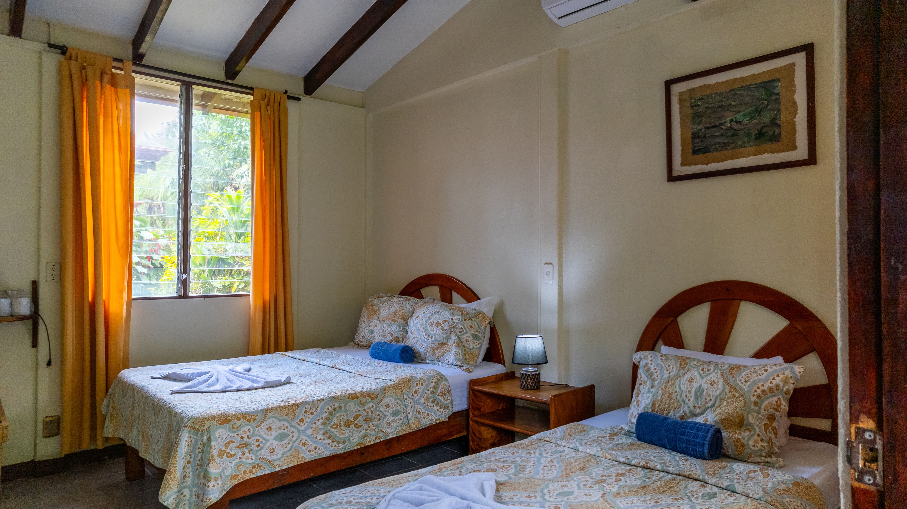 Una habitación con dos camas, cada una con ropa de cama y almohadas estampadas, una mesita de noche con lámpara y una ventana con cortinas.