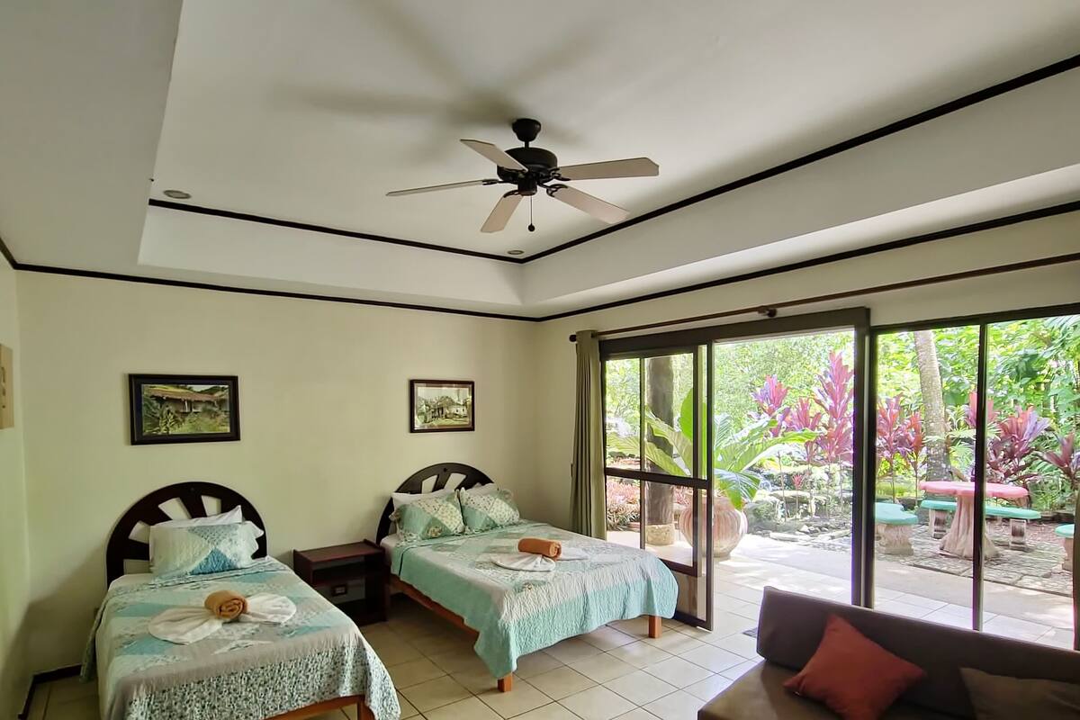 A room with two beds, a sofa, a ceiling fan, and large windows.