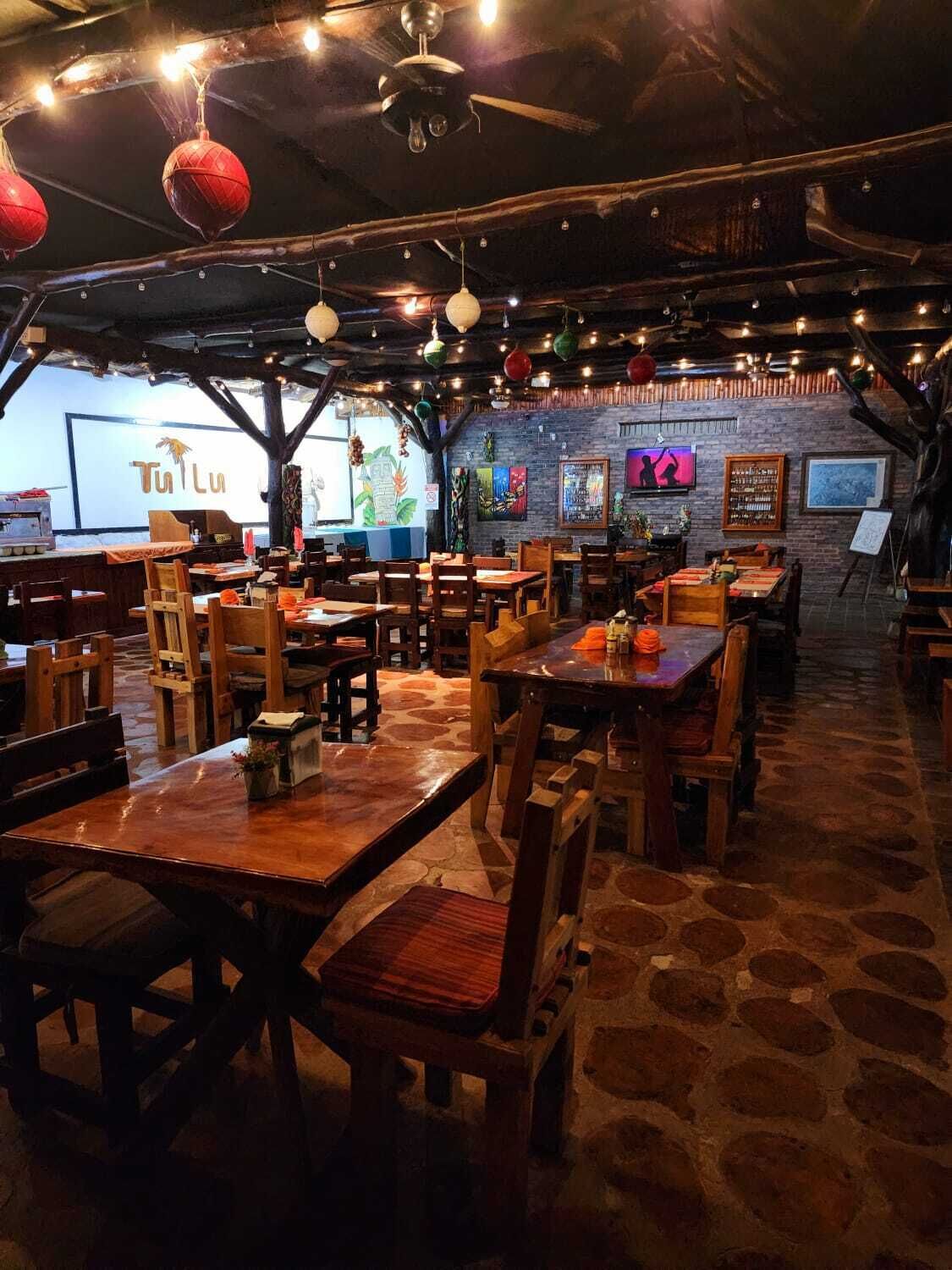 A restaurant with wooden tables and chairs, a brick wall with artwork, and hanging red lanterns.