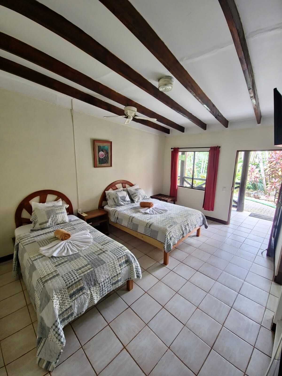 A room with two beds, wooden beams on the ceiling, a ceiling fan, and a window with red curtains.