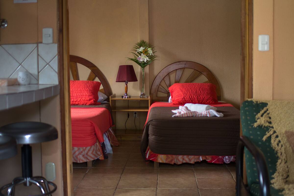 A hotel room with two beds, a small table with a lamp, a vase with flowers, and a chair.