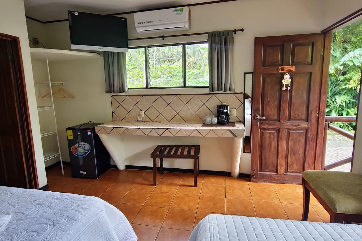 A hotel room with two beds, a kitchenette, a ceiling fan, an air conditioner, and a view of greenery outside.