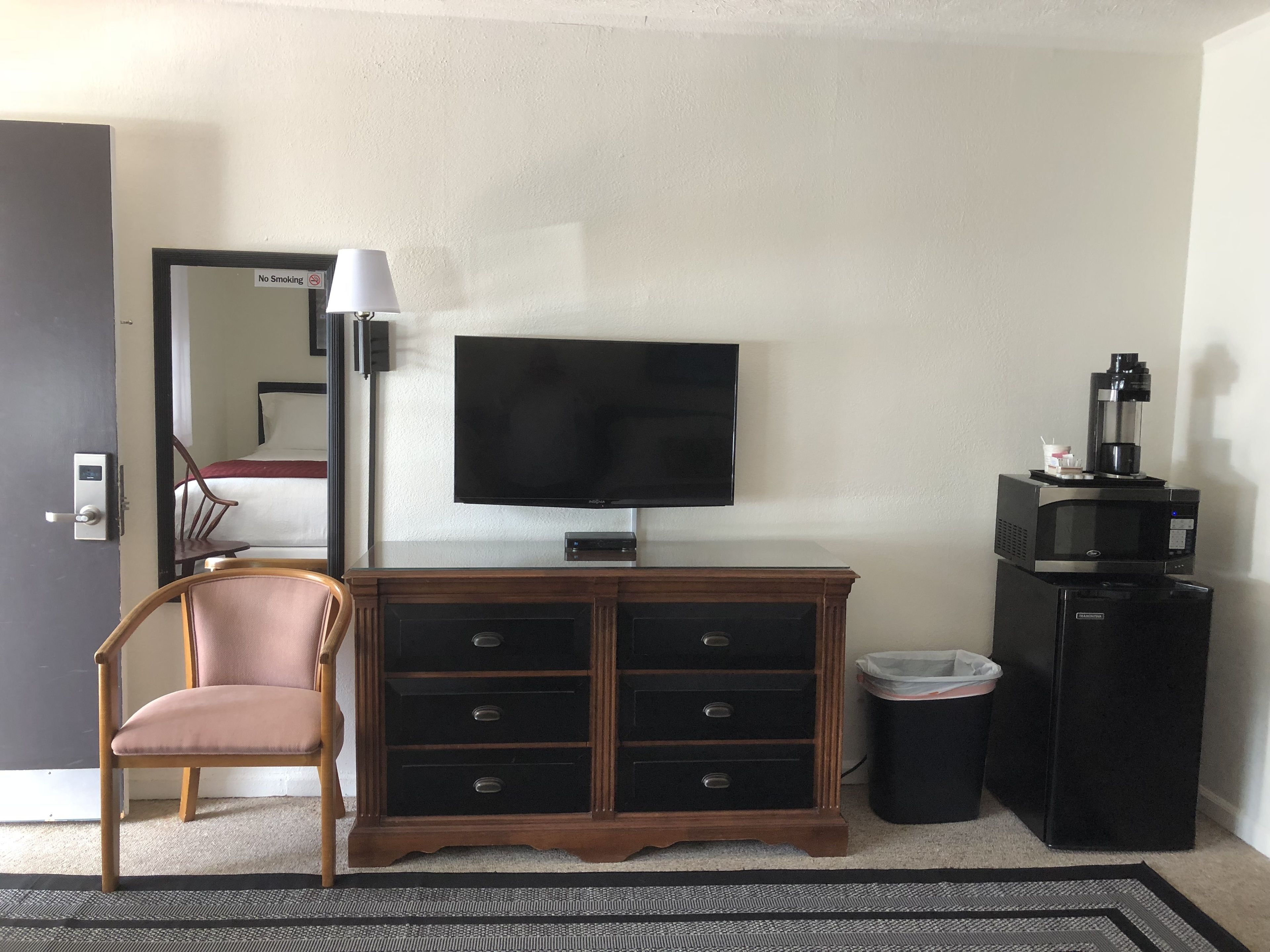 A hotel room with a television, a wooden dresser, a chair, a trash bin, and a coffee machine.
