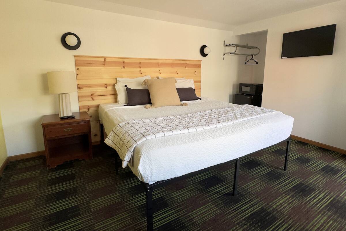 A hotel room with a wooden headboard, a bed with a white comforter, a nightstand with a lamp, a TV on a stand, and a wall-mounted coat rack.