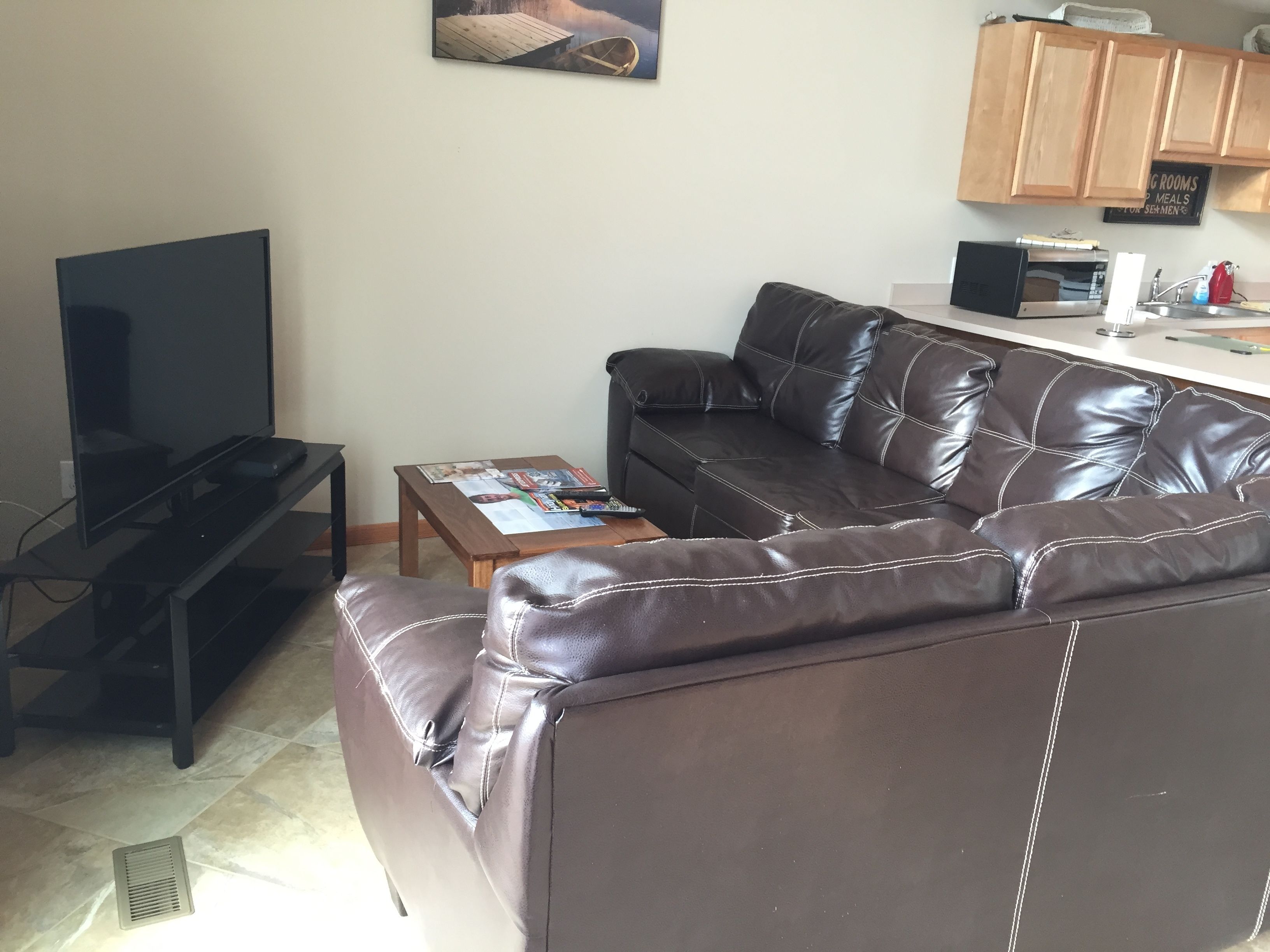 A living room with a brown leather sofa, a coffee table with magazines, a TV on a stand, and wooden cabinets above a countertop.