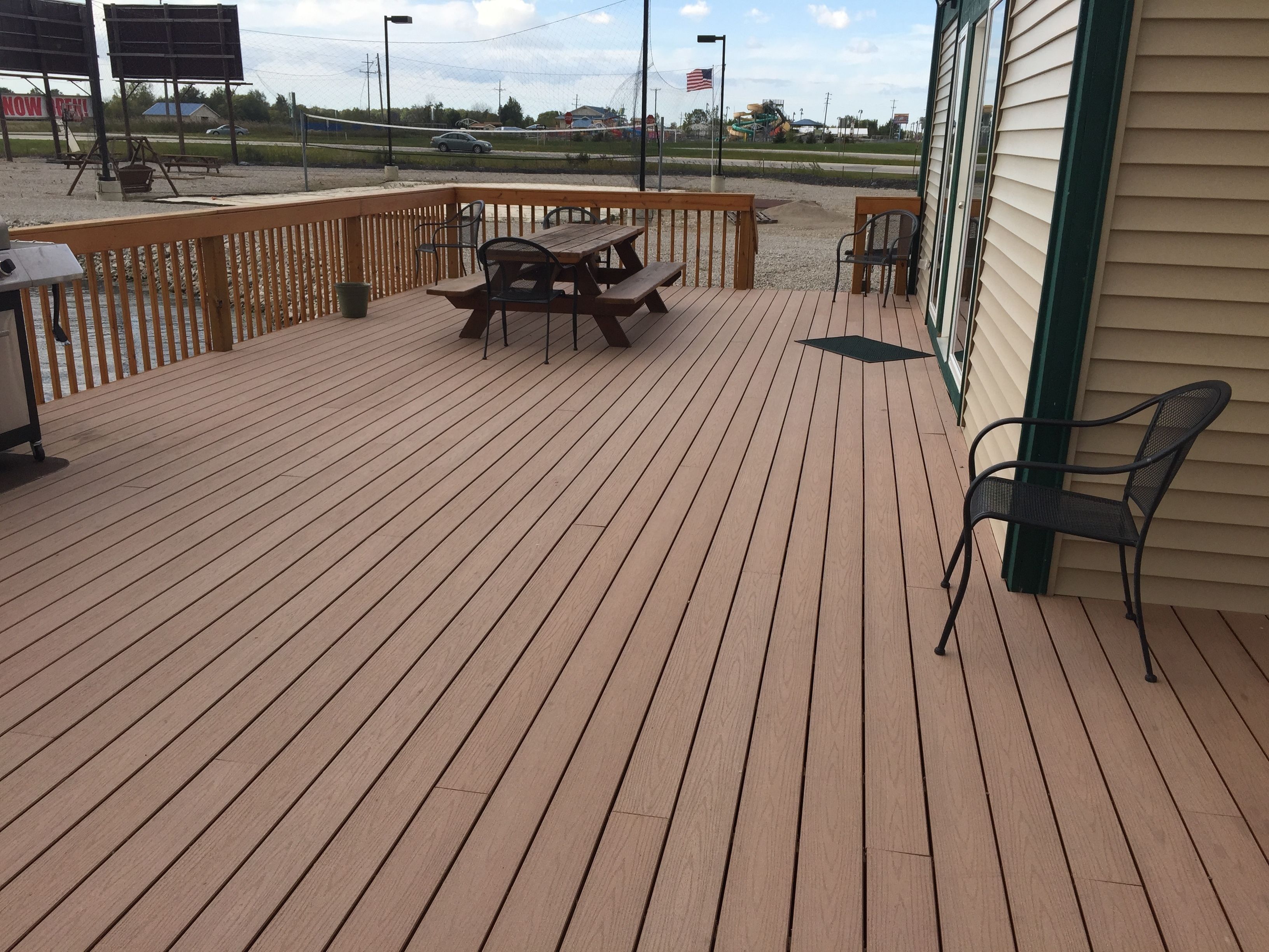 A wooden deck with picnic tables and chairs, a railing, and a view of a playground and cars in the background.