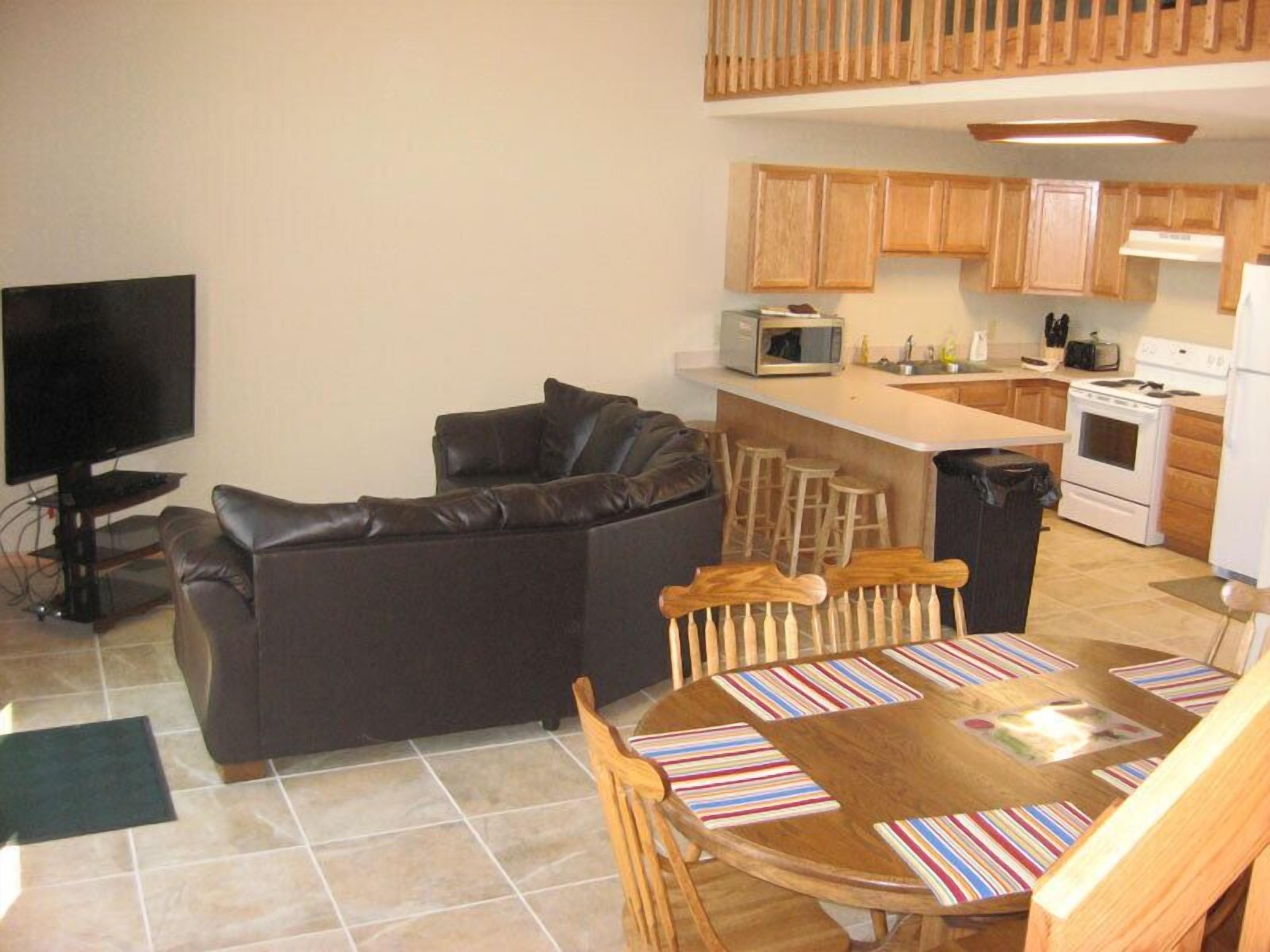A compact living space with a sofa, a dining table with chairs, a kitchen area with wooden cabinets, a microwave, a stove, and a television.