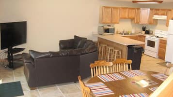A compact living space with a sofa, a dining table with chairs, a kitchen area with wooden cabinets, a microwave, a stove, and a television.