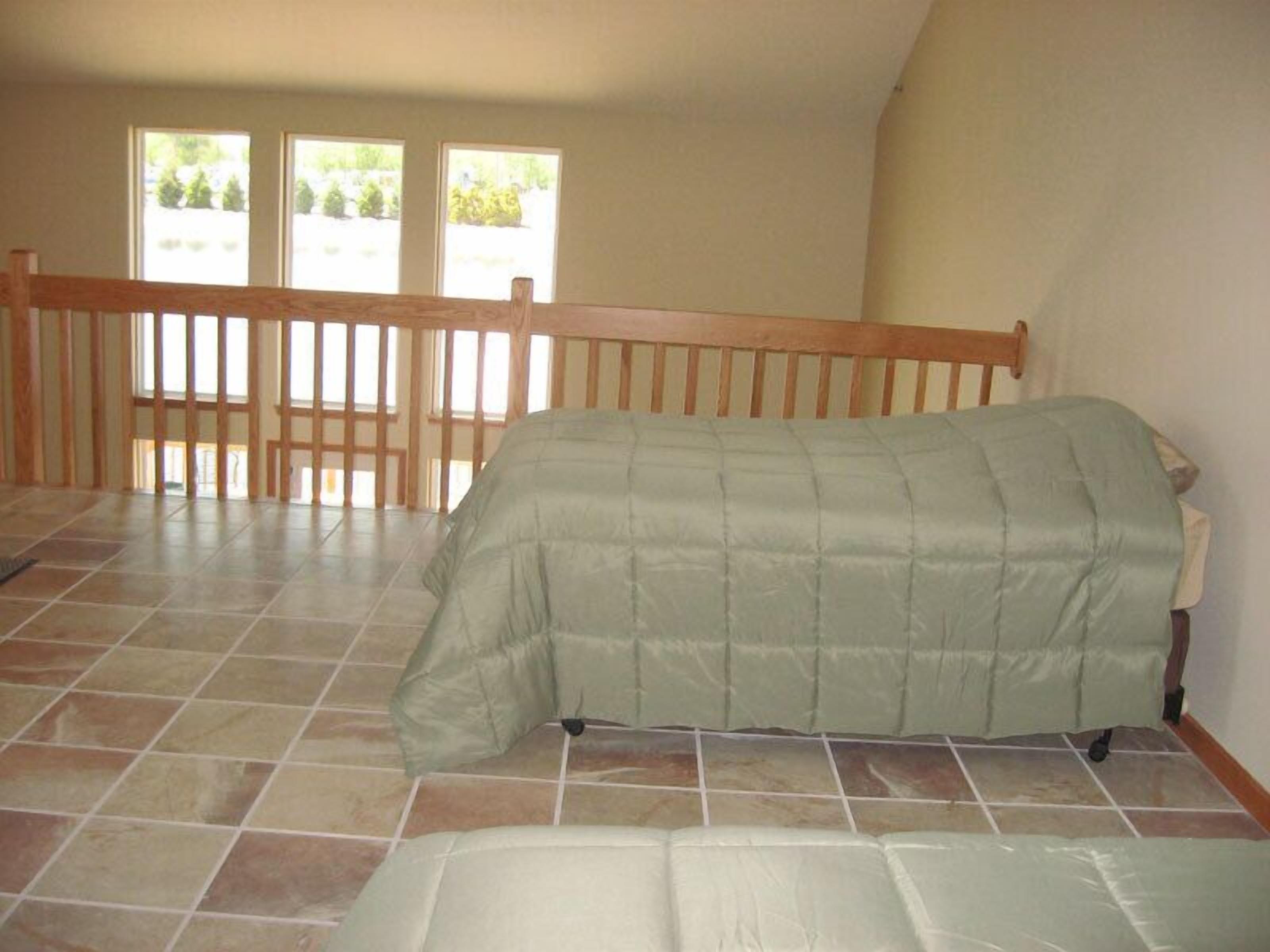 A room with a bed, a wooden railing, and a view of greenery through the windows.