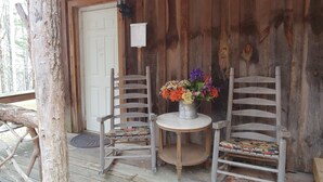 A wooden porch with two rocking chairs and a small table with a flower arrangement. - Arrowmont Stables & Cabins (Cullowhee)