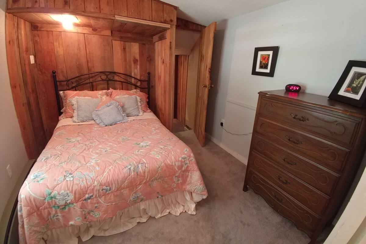 Quarto com cabeceira de madeira, colcha floral, cómoda de madeira e um quadro na parede.