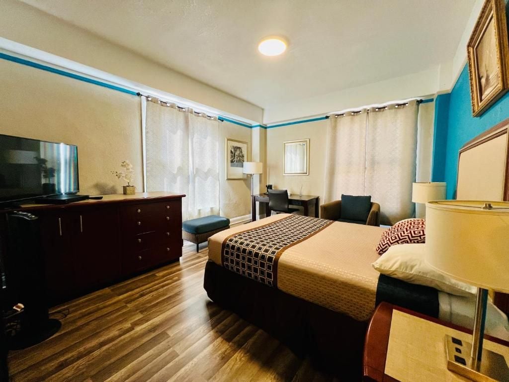 A hotel room with a wooden floor, a bed with a patterned bedspread, a desk with a chair, a television, and a lamp.