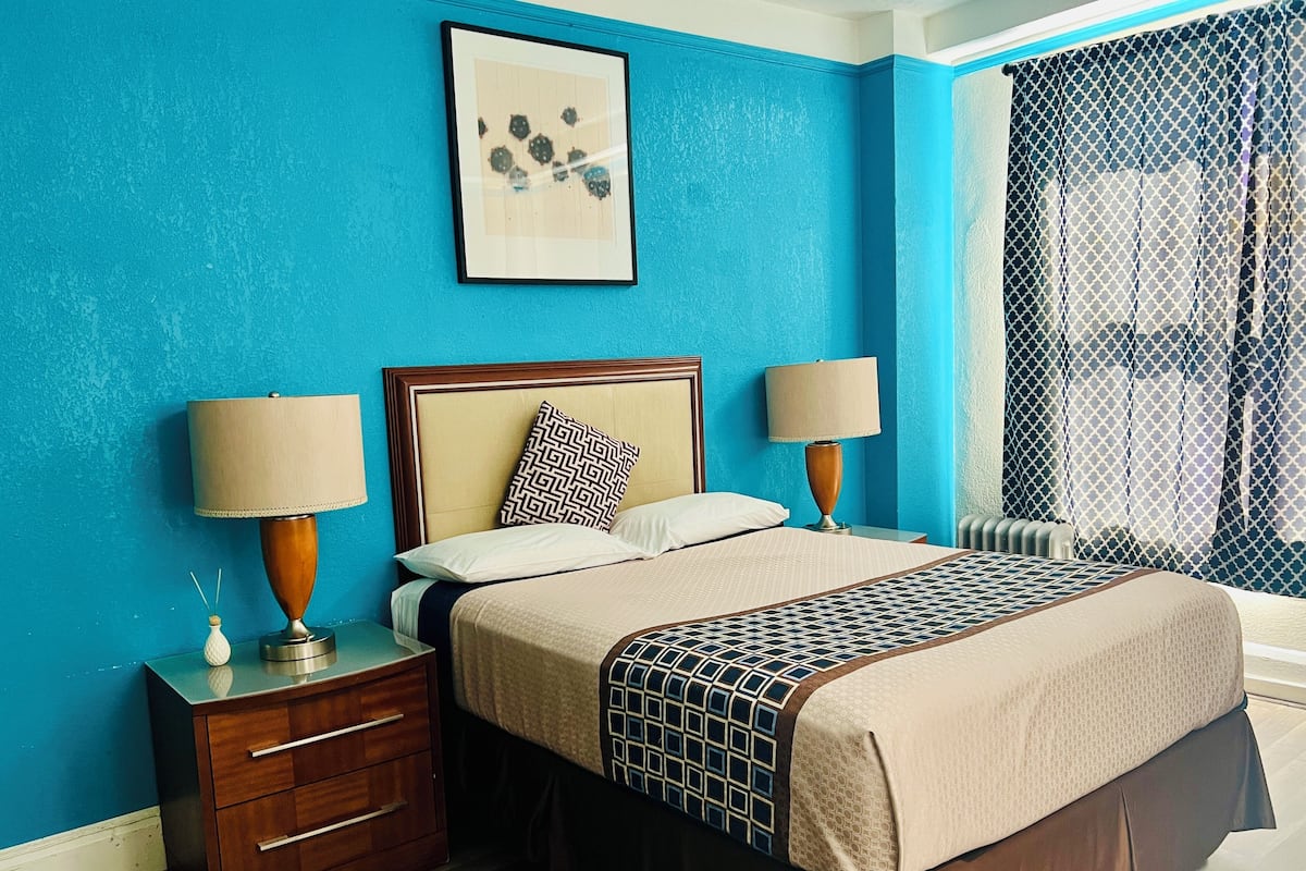 A hotel room with a large bed, two bedside tables with lamps, a nightstand, a framed picture on the wall, and a window with a view.