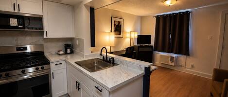 A compact kitchen with a stove, sink, and cabinets, a small dining area with a table and chairs, and a TV in the background.
