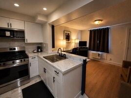 Una cocina compacta con estufa, lavaplatos y armarios, un pequeño comedor con una mesa y sillas, y un televisor al fondo.