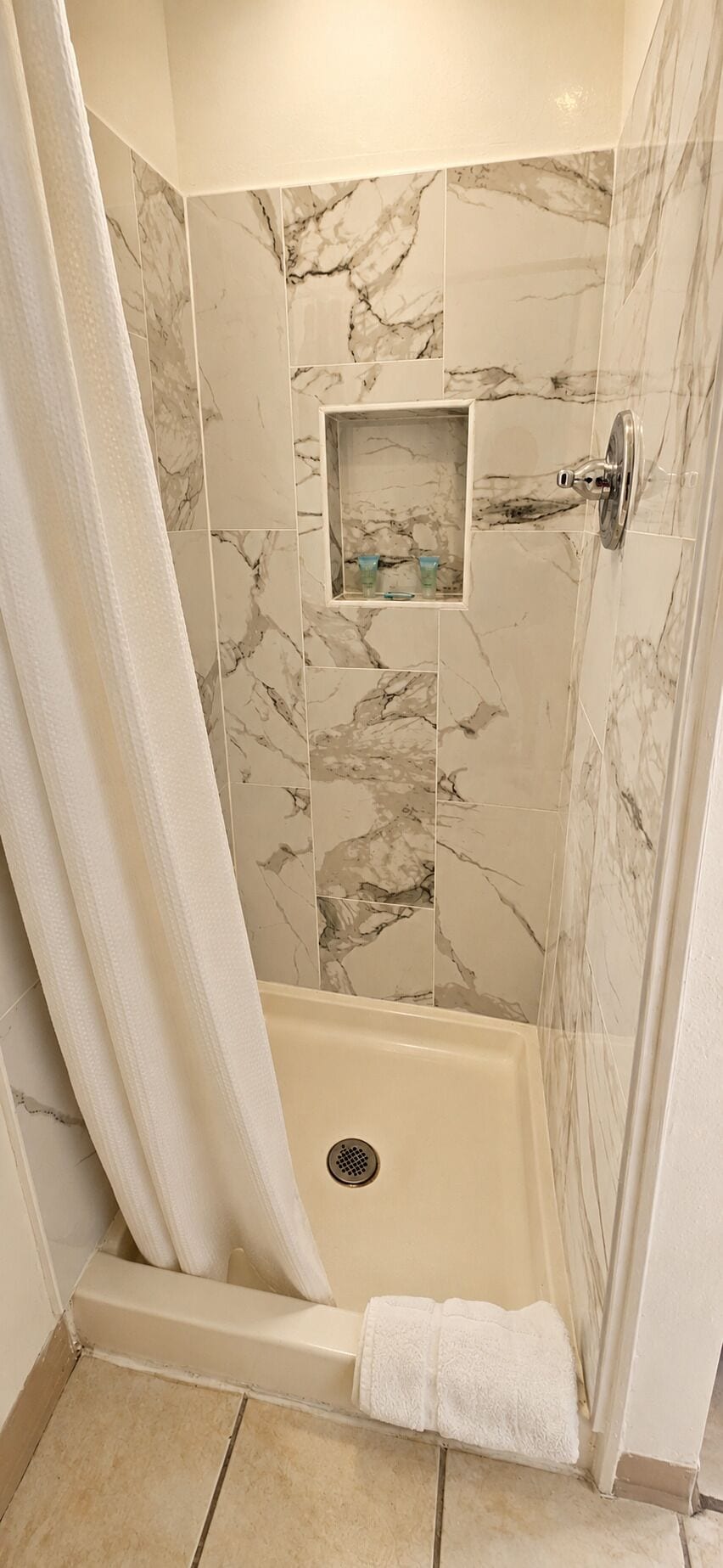 A shower area with a white curtain, a built-in shelf with toiletries, and a white towel on the floor.
