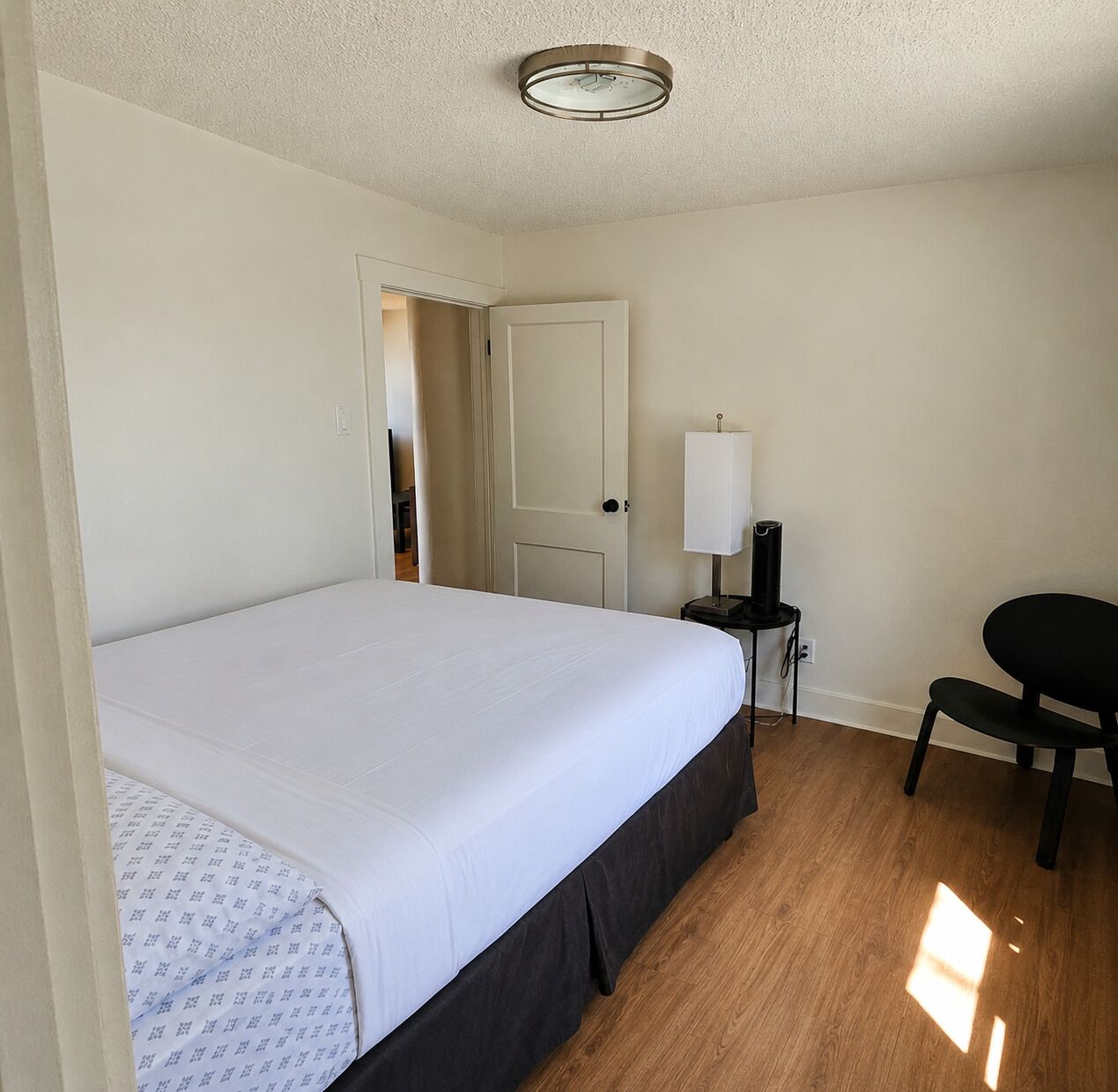 A bedroom with a bed, a desk with a lamp, a chair, and a door leading to another room.