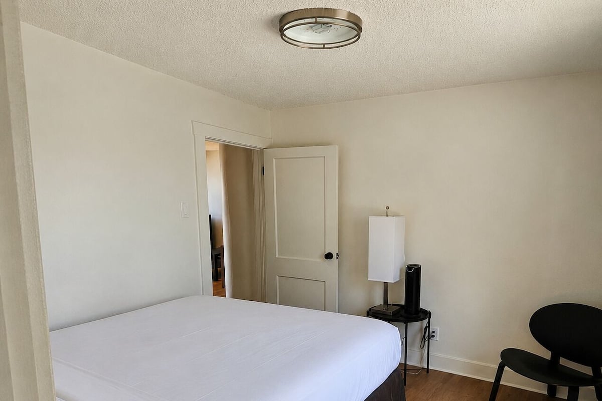 A bedroom with a bed, a desk with a lamp, a chair, and a door leading to another room.