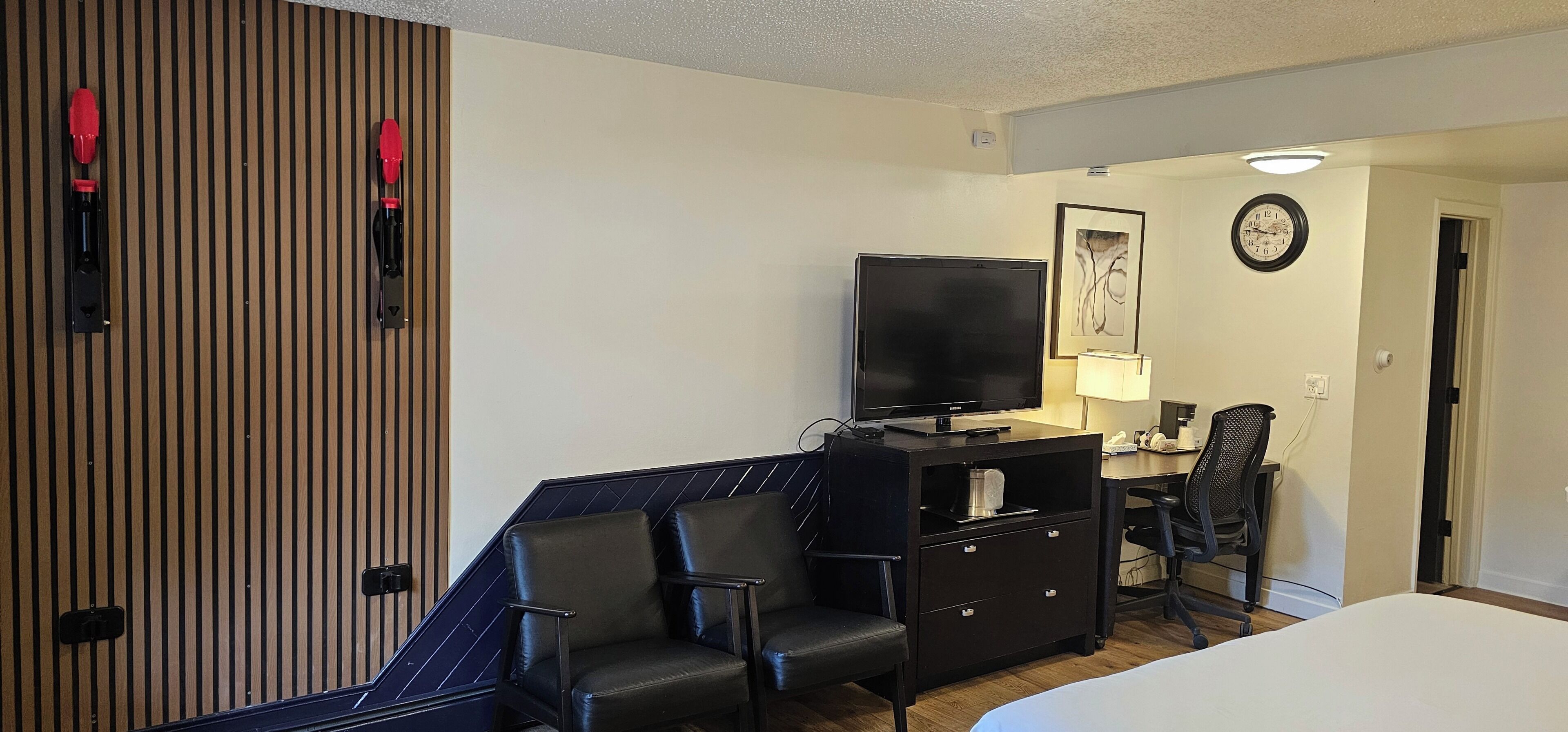 Une chambre d’hôtel comprenant un lit, un bureau avec une télévision, deux chaises, une commode et une horloge fixée au mur.