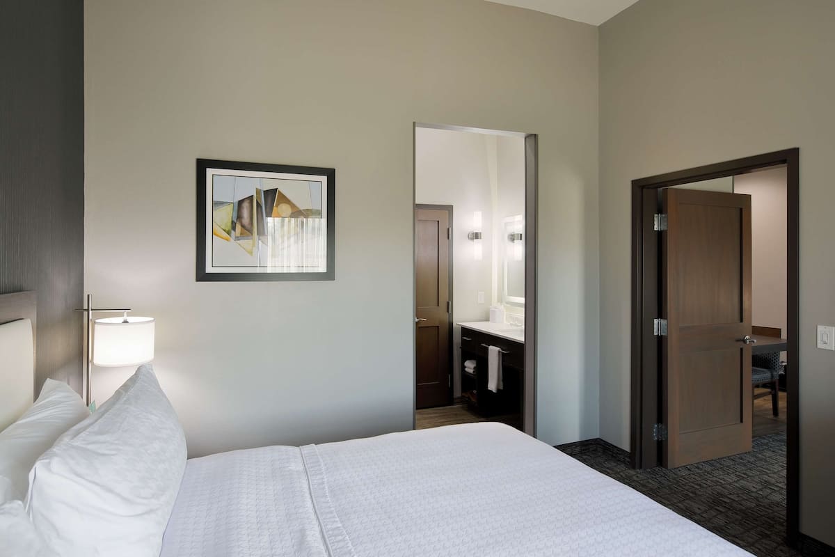 A hotel room with a bed, a bedside lamp, a framed picture on the wall, a bathroom visible through an open door, and a mirror.