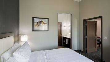 A hotel room with a bed, a bedside lamp, a framed picture on the wall, a bathroom visible through an open door, and a mirror.
