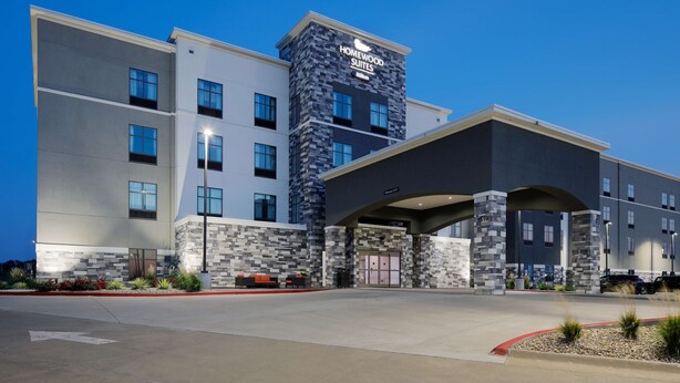Un hôtel au design moderne, reconnaissable à son panneau « Homewood Suites » bien visible, à son entrée couverte et à son espace paysager aménagé avec des sièges.