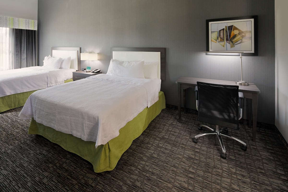 A hotel room with two beds, a desk, a chair, a lamp, and a painting on the wall.