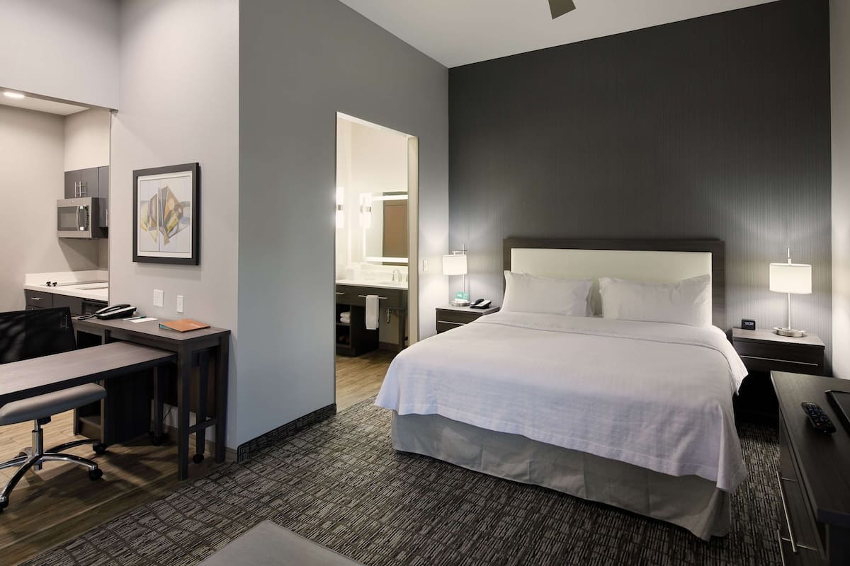 A hotel room with a large bed, a desk, a chair, a desk lamp, a TV, and a bathroom visible through an open door.