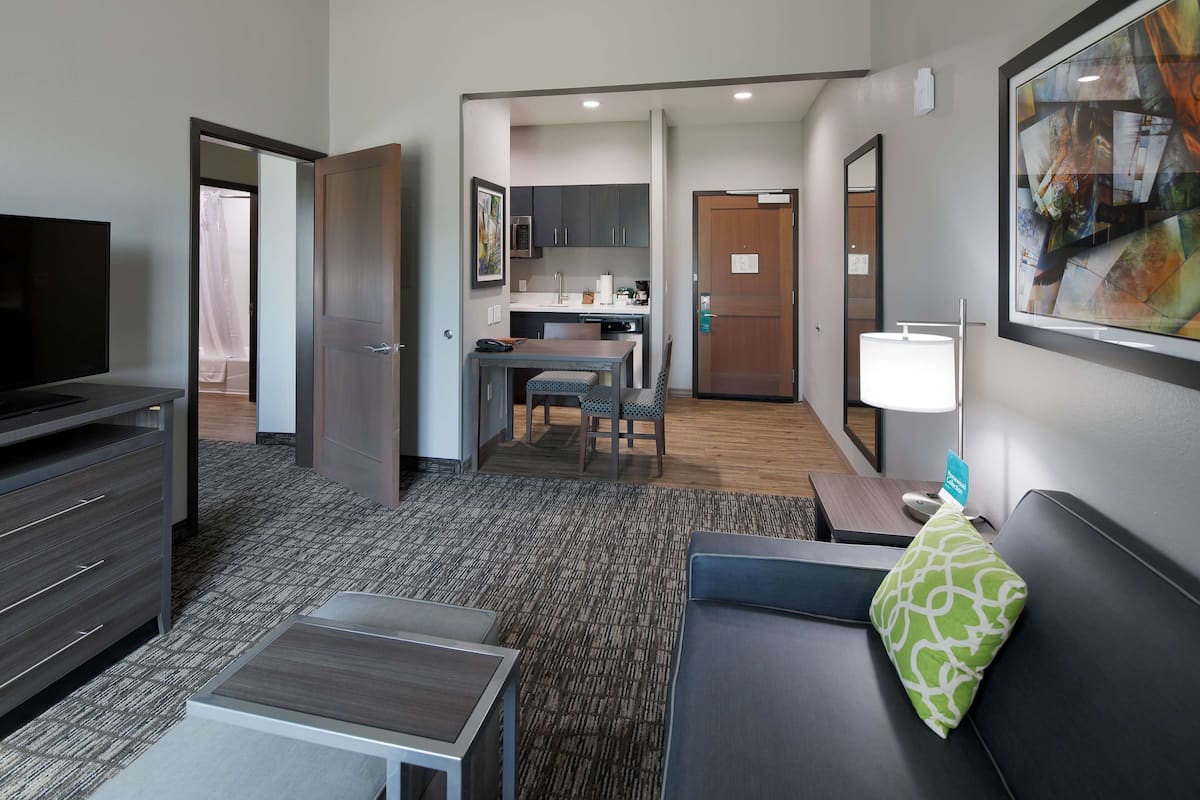 A hotel room with a TV, a sofa, a small table, a kitchen area, and a bathroom.