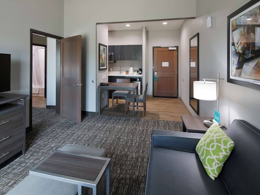 A hotel room with a TV, a sofa, a small table, a kitchen area, and a bathroom.