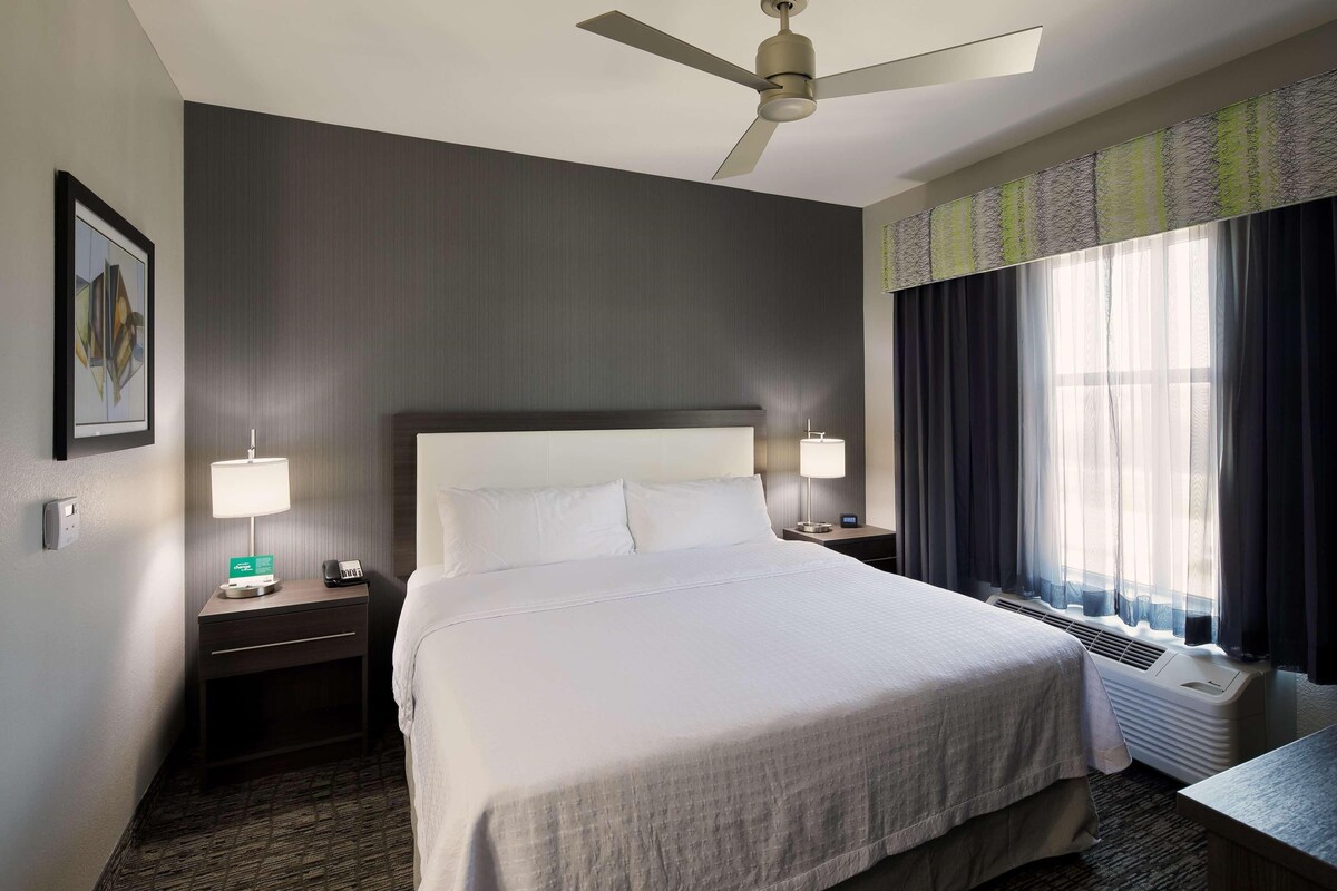 A hotel room with a large bed, two bedside tables with lamps, a ceiling fan, and a window with curtains.