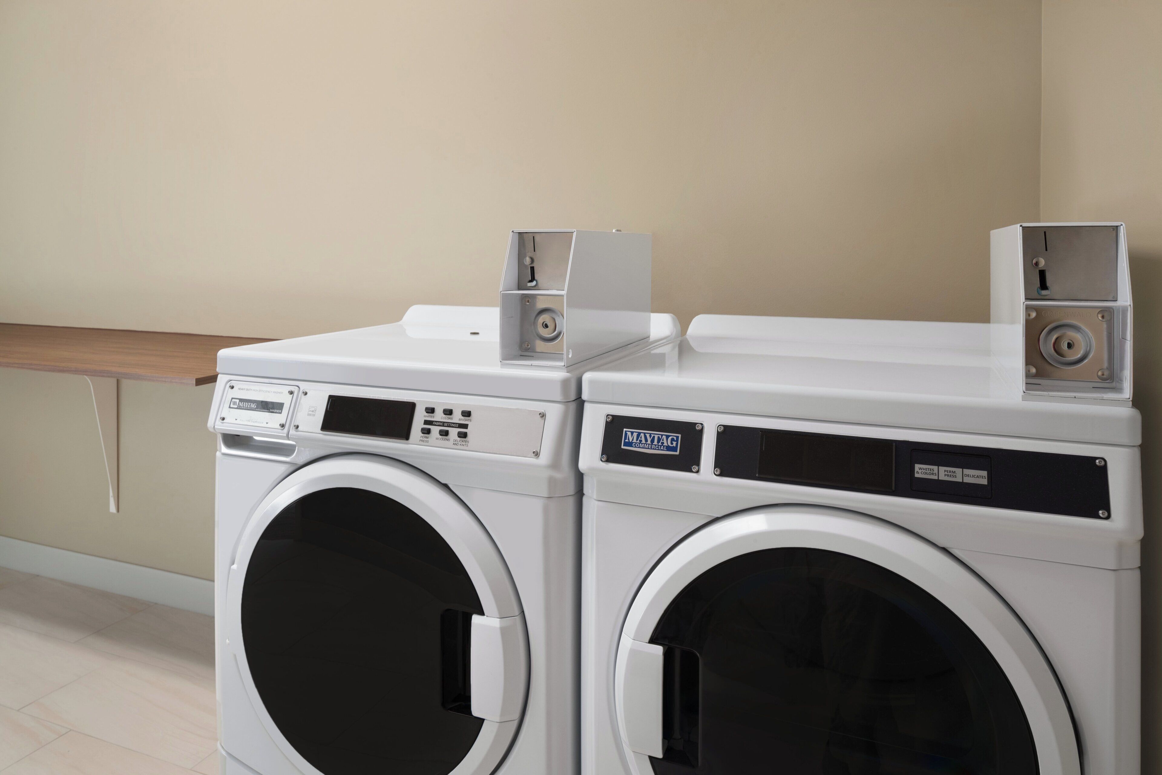 Two front-loading washing machines with control panels, placed next to a wooden bench in a room with a plain wall and tiled floor.