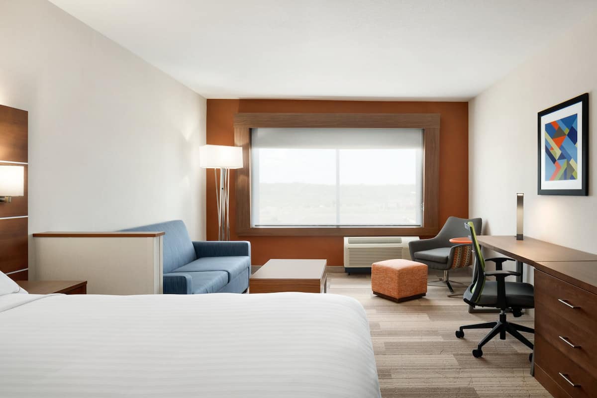 A hotel room with a large bed, a desk, a chair, a sofa, and a window with a view.
