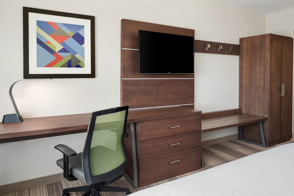 A hotel room with a bed, a desk with a chair, a TV mounted on the wall, and a wardrobe.