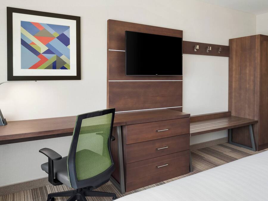 Habitación de hotel con cama, escritorio con silla, televisor montado en un panel de madera y una obra de arte enmarcada en la pared.